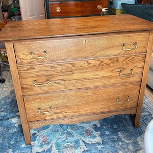 May include: A wooden dresser featuring three horizontal drawers with a natural oak grain finish. Each drawer is equipped with two brass-toned metal handles and a central keyhole escutcheon. The piece stands on four straight legs.