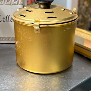 May include: A cylindrical gold-colored metal canister with a matching lid featuring ventilation slits and a black knob handle. The lid is secured to the main body with a small metal latch. The exterior surface shows signs of wear and minor scratches.