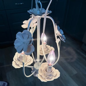 May include: A white floral chandelier with four light bulbs. The chandelier has a central stem with curved arms, each holding a light bulb. The arms are adorned with white and blue flower-shaped accents.