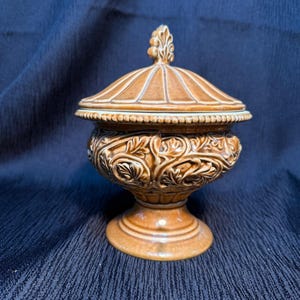 Op de afbeelding: Een bruine keramische pot met deksel en sierlijke bloemenreliëfdetails. De pot heeft een voetstuk en een gewelfd deksel met een decoratieve bekroning. De kleur is een warm, aards bruin.