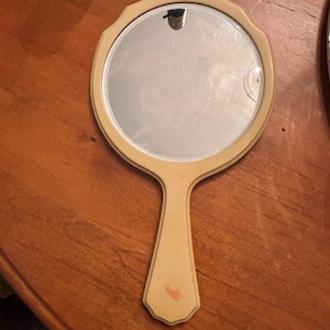 Puede incluir: Un espejo de mano vintage con una superficie de espejo redonda y biselada. El espejo está enmarcado en un color beige claro y tiene un mango a juego. El mango tiene una forma decorativa. El espejo es probablemente de mediados del siglo XX.