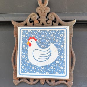 May include: A decorative trivet featuring a white chicken with a red comb on a blue floral background. The ceramic tile is set in a decorative, ornate, cast iron frame. A charming kitchen accent.