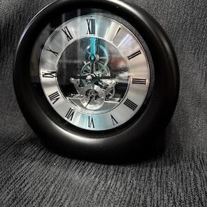 May include: A black, round clock with a silver face and Roman numerals. The clock's inner workings are visible, showcasing the gears and mechanisms. The clock is a decorative item.