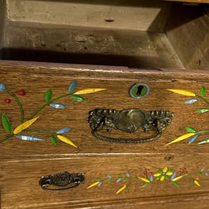 May include: A wooden dresser with an open top drawer featuring a brass handle and a keyhole. The drawer front is decorated with inlaid floral patterns in blue, yellow, green, and red. The wood grain is visible, and the drawer interior is empty.