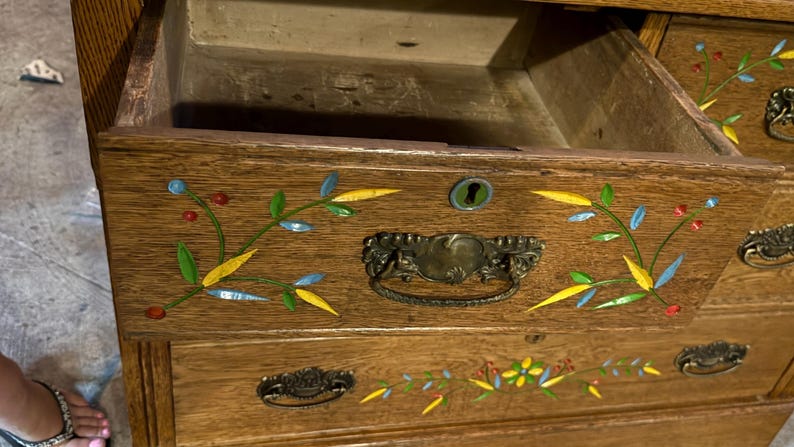 May include: A wooden dresser with an open top drawer featuring a brass handle and a keyhole. The drawer front is decorated with inlaid floral patterns in blue, yellow, green, and red. The wood grain is visible, and the drawer interior is empty.