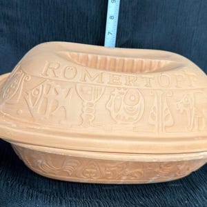 May include: A tan clay Römertopf baking dish with a lid featuring embossed illustrations of fish, goblets, and animals. The lid has the text RÖMERTOPF molded into the surface. A measuring tape is visible, showing a portion of the scale in inches.