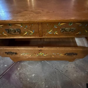 May include: A wooden chest of drawers featuring a natural oak finish with hand-painted floral and bamboo motifs in yellow, blue, and green. The piece includes two top drawers and one larger bottom drawer, all equipped with ornate metal pull handles.