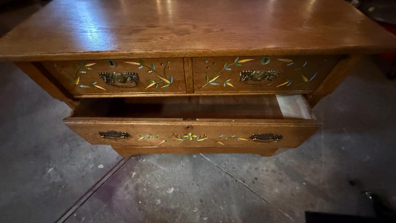 May include: A wooden chest of drawers featuring a natural oak finish with hand-painted floral and bamboo motifs in yellow, blue, and green. The piece includes two top drawers and one larger bottom drawer, all equipped with ornate metal pull handles.