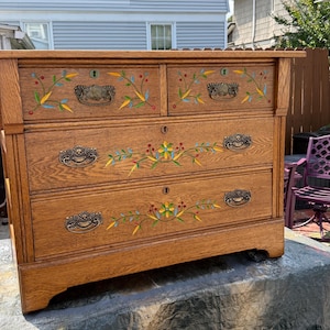May include: A wooden dresser featuring four drawers with ornate metal pulls and keyholes. The front of the drawers is decorated with hand-painted floral motifs in yellow, green, and red, showcasing a natural wood grain finish throughout the piece.