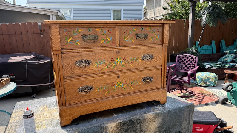 May include: A wooden dresser featuring four drawers with ornate metal pulls and keyholes. The front of the drawers is decorated with hand-painted floral motifs in yellow, green, and red, showcasing a natural wood grain finish throughout the piece.