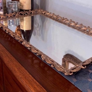 May include: A rectangular, antique-style mirror with a gold-colored, ornate frame. The frame features a repeating leaf design. The mirror is resting on a dark wooden surface, reflecting the objects in front of it.