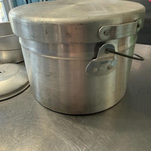 May include: A large, cylindrical, aluminum cooking pot with a lid. The pot has a handle and is sitting on a metal surface. The pot is silver and appears to be used for cooking. The lid sits on top of the pot.