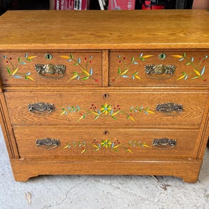 May include: A wooden dresser with a natural oak finish featuring two small top drawers and two large bottom drawers. Each drawer is adorned with hand-painted floral motifs in yellow, green, and red, and includes ornate brass metal drawer pulls.