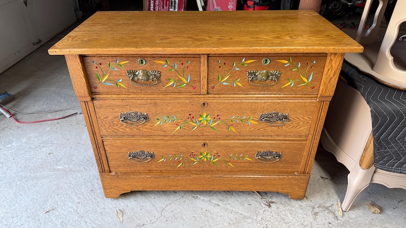 May include: A wooden dresser with a natural oak finish featuring two small top drawers and two large bottom drawers. Each drawer is adorned with hand-painted floral motifs in yellow, green, and red, and includes ornate brass metal drawer pulls.