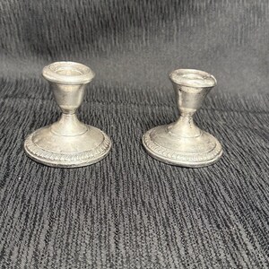 May include: A pair of silver-colored metal candle holders with a polished finish. Each holder features a wide, circular base with a decorative rope-like border, a slender tapered stem, and a flared top opening designed to hold a single candle.