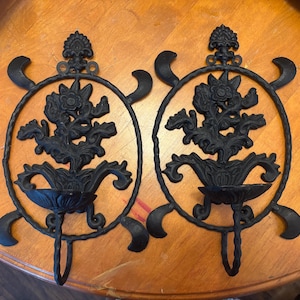May include: Two black metal wall sconces with floral and scrollwork designs. Each sconce features a circular frame with decorative elements, a central floral arrangement, and a small cup-shaped holder for a candle. The sconces are mounted on a wooden surface.
