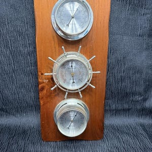 Puede incluir: Una estación meteorológica de madera con tres instrumentos con esferas plateadas. Los instrumentos superior e inferior son circulares, mientras que el del medio tiene forma de timón. La madera tiene una veta natural y un tono marrón cálido. Los instrumentos miden la presión atmosférica.