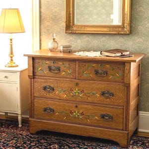 May include: A wooden dresser featuring four drawers with dark metal handles and hand-painted floral motifs in green, yellow, and red. The piece has a natural wood grain finish, keyholes on each drawer, and a rectangular frame structure.