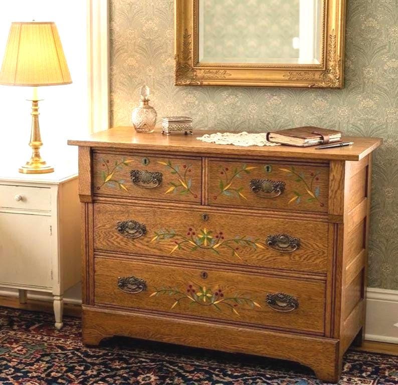 May include: A wooden dresser featuring four drawers with dark metal handles and hand-painted floral motifs in green, yellow, and red. The piece has a natural wood grain finish, keyholes on each drawer, and a rectangular frame structure.