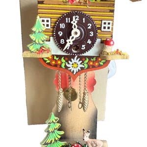 Pode incluir: Um relógio de cuco de madeira com telhado vermelho, mostrador castanho com números brancos e elementos decorativos, incluindo uma árvore verde, um cogumelo vermelho e um pequeno veado. Três pesos dourados em forma de pinha pendem de correntes.