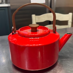May include: A bright red teapot with a cylindrical body, a matching red lid, and a brown wooden handle that arches over the top. The lid features a small, round brown wooden knob in the centre, and the spout extends from the side of the vessel.