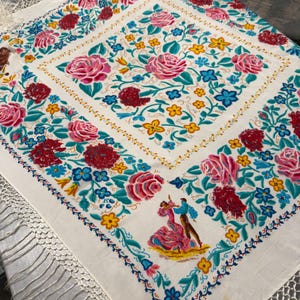 Puede incluir: Un mantón cuadrado de color blanco roto con un patrón floral central de rosas rosas y pequeñas flores azules y amarillas. El borde muestra rosas más grandes, claveles y una pequeña ilustración de una pareja bailando. Flecos blancos largos.