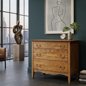 May include: A three-drawer wooden dresser with brass hardware. The dresser is made of light-colored wood and has a classic design. A gold-colored sculpture and a framed line art print are visible in the background. A small gold clock and a vase with a plant are on top of the dresser.