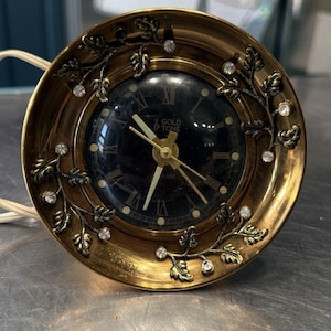 May include: A vintage gold-toned alarm clock with a black face and Roman numerals. The clock features decorative leaf and crystal accents around the bezel. The text "GOLD TONE" is visible on the clock face. A white cord extends from the clock.