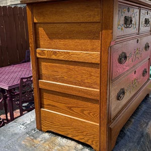May include: A wooden dresser with a medium brown finish, featuring four drawers with dark metal hardware. Each drawer front is decorated with hand-painted floral motifs in green, yellow, and red. The side panel shows horizontal wood grain detailing.