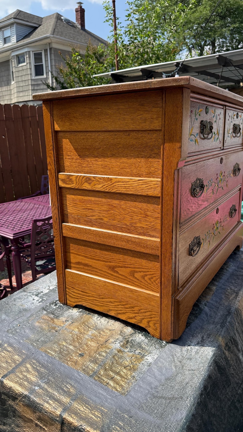 May include: A wooden dresser with a medium brown finish, featuring four drawers with dark metal hardware. Each drawer front is decorated with hand-painted floral motifs in green, yellow, and red. The side panel shows horizontal wood grain detailing.