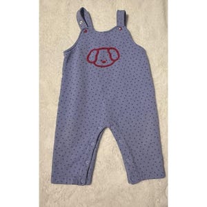May include: A baby's blue overall with a repeating dark blue polka dot pattern. The front features a red embroidered dog face. The overall has snap closures on the legs and shoulder straps.