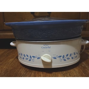 Puede incluir: Una olla de cocción lenta Rival Crock-Pot blanca con un inserto de cerámica azul y tapa de vidrio. La base presenta un patrón de enredadera con hojas azules, un mando central con ajustes Off, Low, High, Warm y dos asas laterales.