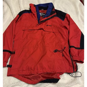 May include: A red Tyrolia anorak jacket with black and blue accents. The jacket features a high collar, snap closures, and a drawstring at the waist. Black stripes are on the sleeves. The jacket has two front pockets and a zipper on the side.