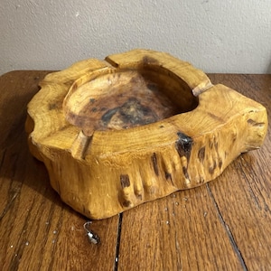 Puede incluir: Un cenicero de madera artesanal con forma irregular. Tallado de un tronco de árbol, presenta un cuenco central y ranuras para cigarrillos. La madera tiene un tono marrón dorado cálido con nudos y vetas más oscuras.