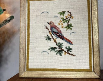 May include: A framed needlepoint artwork featuring a colorful bird perched on a branch with green leaves and yellow berries. The background is a cream color with blue and white stitching.