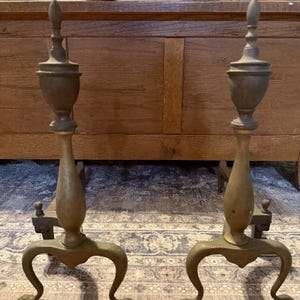 May include: Two antique brass and bronze fireplace andirons. Each has an urn-shaped top, a slender stem, and a curved base. The andirons are set against a wooden background. The andirons are a decorative fireplace accessory.