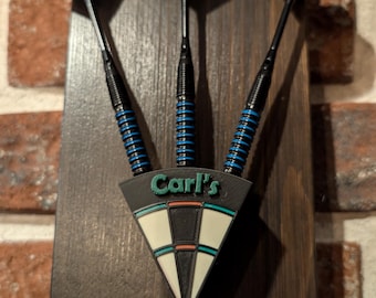 Puede incluir: Un soporte de dardos de madera con forma triangular y el texto "Carl's" impreso en él. Tres dardos negros y azules están unidos al soporte.