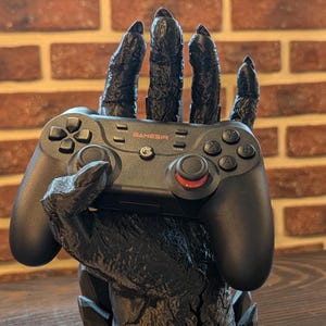 Könnte beinhalten: Ein schwarzer GAMESIR-Controller ruht in einem schwarzen, stilisierten handförmigen Ständer. Der Ständer hat ein strukturiertes, raues Aussehen, mit sichtbaren Knöpfen und roten Akzenten des Controllers. Der Hintergrund zeigt eine Backsteinmauer.
