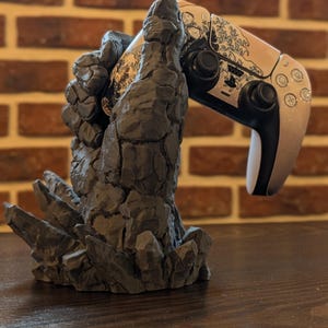 Può includere: Un supporto per controller grigio, simile a una roccia, che tiene in mano un controller per videogiochi argento e nero. Il controller ha un design decorativo sulla parte superiore. Il supporto è su una superficie di legno scuro con uno sfondo di mattoni.