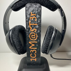 Könnte beinhalten: Schwarze Kopfhörer mit einem schwarzen Ständer. Der Ständer hat einen schwarz-weißen Aufkleber mit dem Text "1C3M@ST3R" in orange.