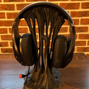Può includere: Cuffie da gioco nere appoggiate su un supporto nero e astratto per cuffie. Il supporto ha un design organico con aperture. Le cuffie hanno grandi padiglioni auricolari e un microfono con punta rossa. Lo sfondo è un muro di mattoni. Il supporto è alto circa 20 cm.