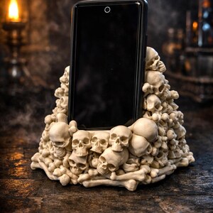 Könnte beinhalten: Ein schwarzes Smartphone ruht in einem beigefarbenen Handyhalter mit Totenkopfmotiv. Der Halter besteht aus zahlreichen Totenköpfen und Knochen und erzeugt eine gotische oder Halloween-Ästhetik. Der Hintergrund ist dunkel und atmosphärisch.