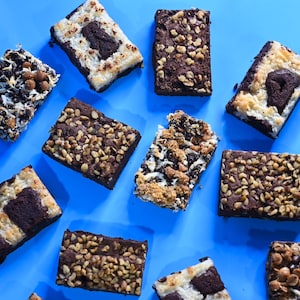 May include: Assortment of brownies with different toppings, including nuts, marshmallows, and chocolate chips. The brownies are arranged on a blue surface.