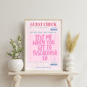 May include: A framed pink "Guest Check" print with the text "TEXT ME WHEN YOU GET TO TUSCALOOSA XO". The print has a vintage aesthetic, with a grid pattern and the words "Thank you. Please come again." at the bottom.