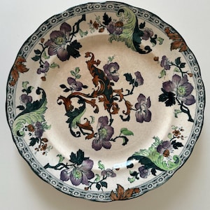 May include: A round ceramic plate with a scalloped edge featuring a cream-colored base. The surface is decorated with a central brown scrollwork design surrounded by purple flowers, dark green leaves, and light green foliage accents.