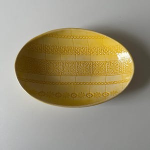 May include: An oval-shaped yellow ceramic bowl featuring a textured surface with horizontal bands of intricate lace-like patterns. The design includes repeating geometric shapes and floral motifs embossed into the glaze, creating a decorative relief.