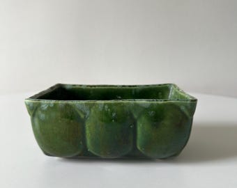 May include: A rectangular ceramic bowl with a glossy, deep green glaze. The exterior features a repeating molded pattern of rounded, semi-circular segments along the sides, creating a textured appearance with subtle variations in color depth.