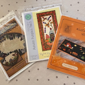 May include: Three rectangular quilt pattern booklets in clear plastic sleeves. The left booklet is titled Primitive Gatherings The Neighborhood. The center booklet is Nicole's Autumn Pumpkin Patch. The right booklet is titled Nutmeg Hare Pumpkin Farm.