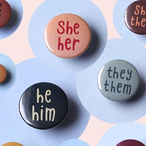 Könnte beinhalten: Eine Sammlung runder Pronomen-Buttons in verschiedenen Farben, darunter Braun, Pfirsich und Grau. Jeder Button zeigt Pronomen wie "She/Her", "He/Him" und "They/Them" sowie deutsche und englische Pronomenoptionen. Die Buttons sind auf einem hellblauen Hintergrund angeordnet.