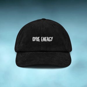 Peut inclure: Casquette de baseball noire en velours côtelé avec le texte blanc "DYKE ENERGY" brodé sur le devant. La casquette a une visière incurvée et une couronne arrondie. Le fond est un dégradé de bleu.